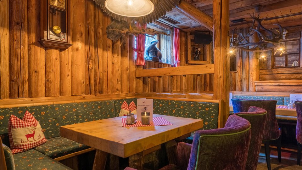 Eine gemütliche Stube mit Holzpaneelen und bequemen Sitzen. Auf dem Tisch stehen Menükarten und die Atmosphäre ist einladend.