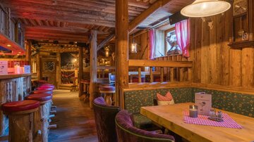 Eine gemütliche Hütte mit Holzinterieur und warmem Licht. Es gibt Tische und bunte Sitzgelegenheiten, die einladend wirken.