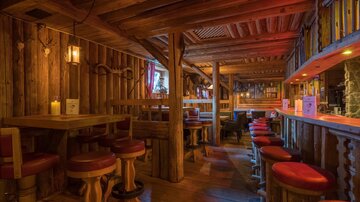Eine gemütliche Bar mit Holzverkleidung und warmem Licht. Die Einrichtung besteht aus Tischen und Stühlen mit roten Polstern.