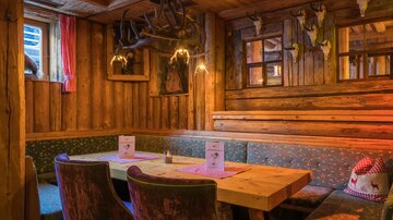 Ein rustikales Holzinterieur mit einem großen Tisch und bequemen Sitzplätzen. Die Wände sind mit Geweihen dekoriert, und die Beleuchtung schafft eine gemütliche Atmosphäre.