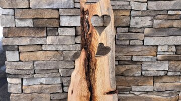 Ein künstlerisches Holzobjekt mit Herzformen und einer rustikalen Optik. Es steht vor einer steinernen Wand und vermittelt eine gemütliche Atmosphäre.