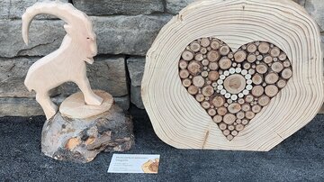 Ein handgefertigtes Holzskulpturen-Set. Es zeigt eine Ziegenfigur und ein Herz aus Holzscheiben auf einer Steinmauer.