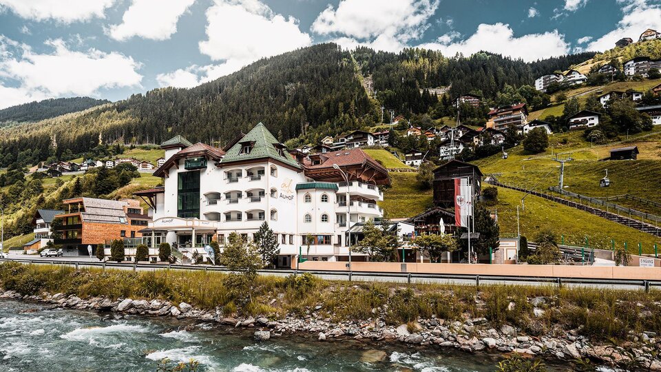 Ein malerisches Hotel am Flussufer, umgeben von grünen Hügeln und Bergen. Die Gebäude sind von schönem, ländlichem Flair geprägt.