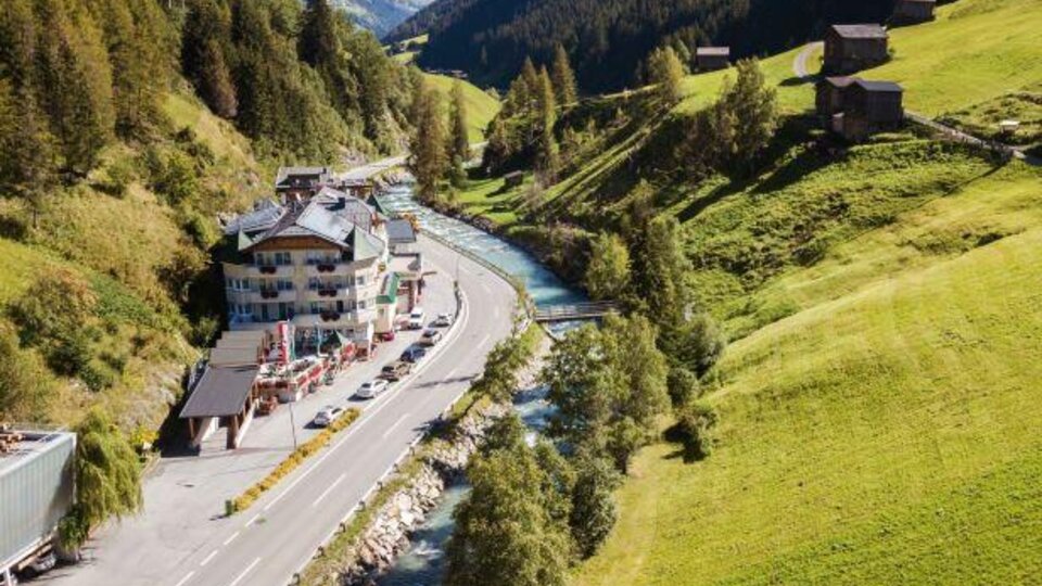 Eine malerische Landschaft mit einem Fluss, der durch ein Tal fließt. Am Ufer stehen ein paar Häuser und die Umgebung ist von grünen Wiesen und bewaldeten Hügeln umgeben.