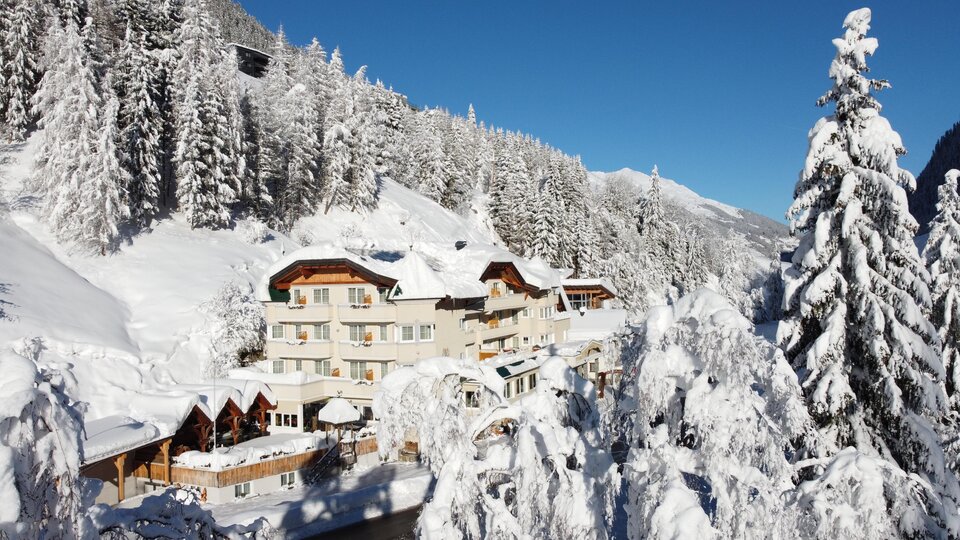 Ein malerisches Hotel in einer schneebedeckten Winterlandschaft. Hohe Tannenbäume und ein klarer blauer Himmel umgeben die Szene.