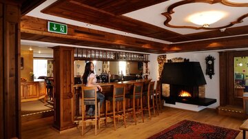 Eine gemütliche Bar mit Holzverkleidung und einem offenen Kamin. Im Hintergrund steht eine Frau an der Bar und es gibt mehrere Sitzplätze.