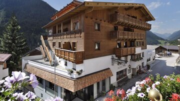 Ein charmantes Holzgebäude mit mehreren Balkonen und blühenden Blumen am Eingang. Im Hintergrund sind malerische Berge und ein klarer Himmel zu sehen.