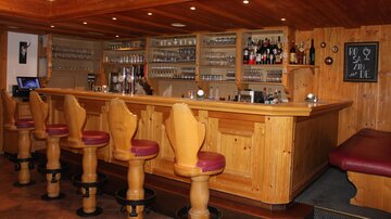 Eine gemütliche Bar mit Holzverkleidung und Hochstühlen. Hinter der Bar stehen verschiedene alkoholische Getränke und Gläser.
