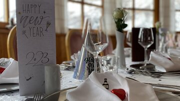 Ein festlich gedeckter Tisch für Silvester mit eleganten Tellern, Gläsern und dekorierten Servietten. Im Hintergrund steht eine Tafel mit der Aufschrift "Happy New Year 2023".
