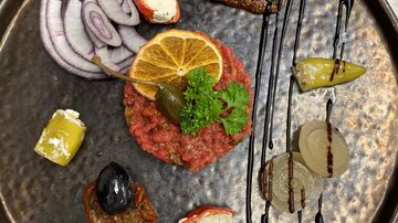 Ein kunstvoll angerichtetes Beefsteak Tatar mit Zwiebeln, Zitronenscheibe und verschiedenen Beilagen. Auf dem Teller befinden sich auch schwarze Oliven und eine raffinierte Sauce.