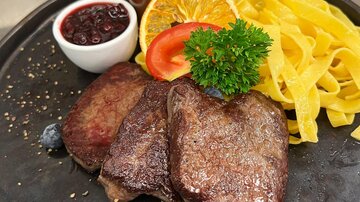Ein elegant angerichteter Teller mit saftigem Steak, frischen Bandnudeln und Beilagen aus Orange und Tomate. Dazu gibt es eine kleine Schüssel mit Preiselbeeren und eine Sauce.