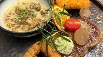 Ein kunstvoll angerichteter Teller mit Fleisch, einer Sauce und verschiedenen Beilagen. Dazu gibt es gefüllte Croquettes, frisches Gemüse und Garnitur.