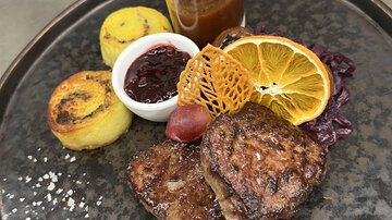 Ein ansprechender Teller mit gegrilltem Fleisch, dekoriert mit einer Orangenscheibe und einer knackigen Süßspeise. Neben einer Portion Sauce und Beilagen aus Polenta und serviertem Obst.