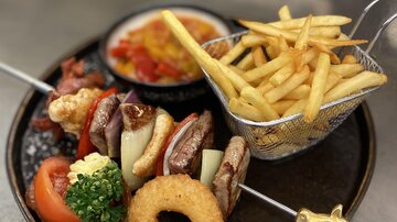 Ein Teller mit gegrilltem Fleisch, Gemüse, einer panierten Zwiebel und Kartoffelspalten. Dazu gibt es Pommes frites und eine scharfe Sauce.