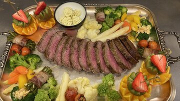 Ein kunstvoll angerichteter Fleischteller mit perfekt gegartem Rindersteak, umgeben von buntem Gemüse und frischen Früchten. In der Mitte befindet sich eine Schüssel mit Sauce.
