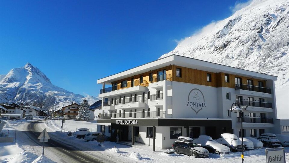 A modern hotel named "Zontaja" in a winter mountain landscape. The surroundings are covered in snow, and majestic mountains can be seen in the background.