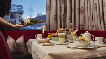 A set table with various breakfast dishes and a glass of sparkling wine. In the background, windows with a view of the mountains can be seen.