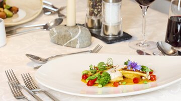 An elegantly arranged dish on a large white plate. In the background, there are cutlery, a candle, and a glass of red wine.