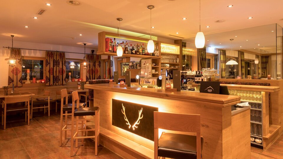 An inviting bar with wooden furniture and dim lighting. In the background, counters and shelves stocked with drinks can be seen.
