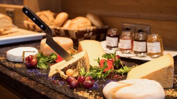 An appealing cheese board with various types of cheese, grapes, and fresh greens. In the background, rolls and jars of jam are visible.