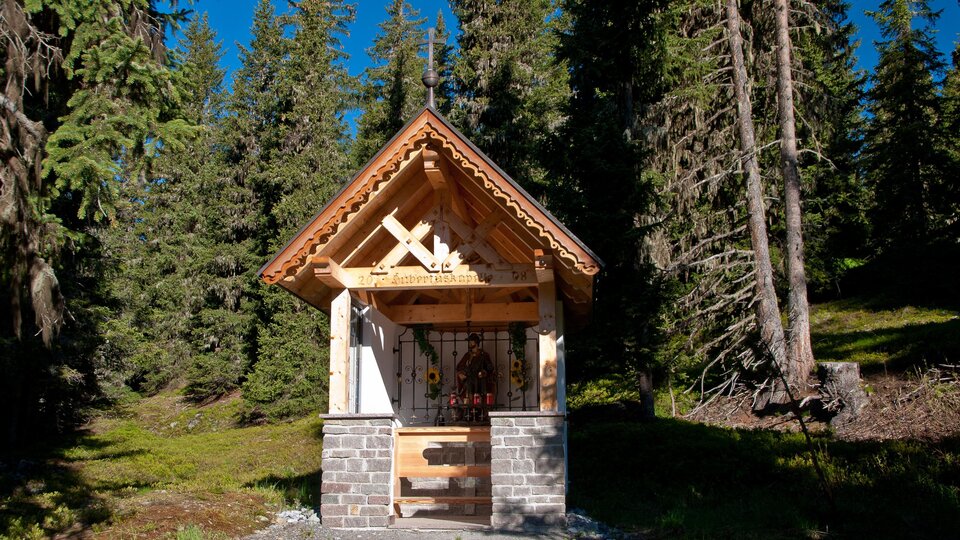 A small wooden chapel surrounded by tall trees. The sky is blue and the surroundings are green and peaceful. | © Bernhard Gruber – Photography