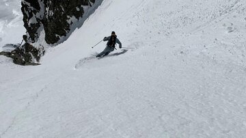 A skier descends the steep, snow-covered slope. In the background, there are rocks and the snowy mountain landscape.