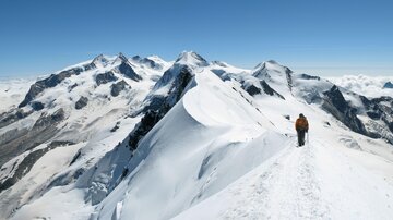 A hiker on a snow-covered peak with impressive mountains in the background. The sky is clear and blue.