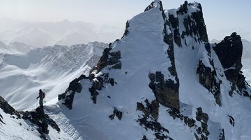 An impressive mountain landscape with snow-covered peaks. In the foreground, a hiker stands on a narrow ridge.