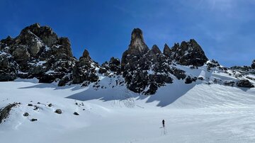 A winter landscape with snow-covered mountains and a clear, blue sky. A person is hiking through the snow.