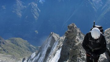 A mountaineer stands on a rocky summit with snow-covered mountains in the background. The sky is clear and blue, and the landscape is majestic.