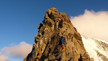 Two climbers ascend a steep rock at sunset. The surroundings are majestic with snow-covered mountains and a clear blue sky.