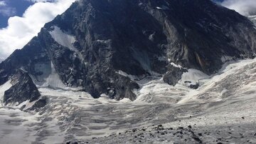 A hiker explores a frozen landscape beneath a majestic mountain. The sky is partly cloudy and conveys a dramatic feeling.