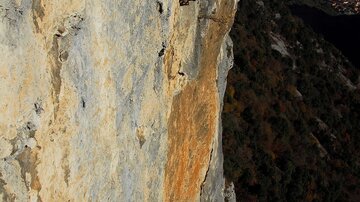 A climber is standing at a steep rock wall. In the background, green mountains and a valley can be seen.