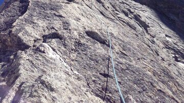 A steep cliff face under a clear blue sky. The sunlight shines on the rocks as climbers begin their ascent.