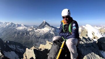 A mountain climber sits on a summit and looks at an impressive mountain landscape. The sun shines brightly on the snow-covered peaks in the background.
