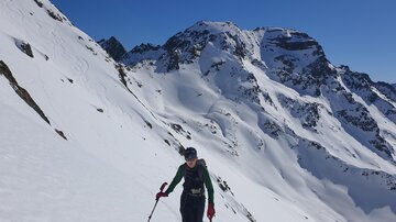 Ein Skifahrer wandert durch eine verschneite Berglandschaft. Im Hintergrund sind majestätische Berge unter einem klaren, blauen Himmel zu sehen.