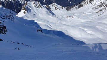 Eine verschneite Berglandschaft mit sanften Hügeln und hochaufragenden Gipfeln. Der Himmel ist klar und die Sonne scheint auf die schneebedeckte Umgebung.