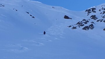 Eine einzelne Person wandert durch eine schneebedeckte Landschaft. Im Hintergrund sind Felsen und sanfte Hügel sichtbar.
