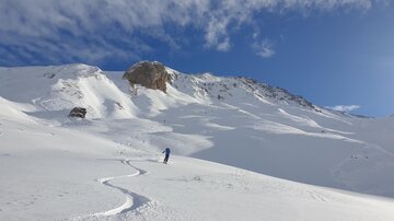 Eine verschneite Berglandschaft mit einem klaren blauen Himmel. Im Vordergrund zieht eine Person Spuren im Schnee.