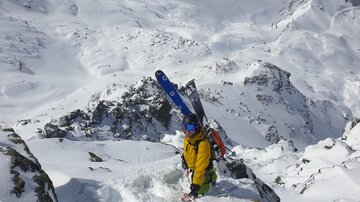 Ein Skifahrer in gelber Jacke steht im Schnee und schaut die geneigte Abfahrt hinunter. Im Hintergrund sind schneebedeckte Berge zu sehen.