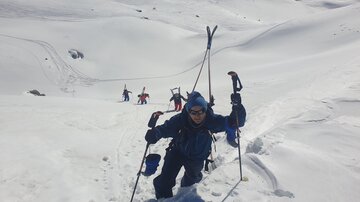 Ein Skialpinist besteigt einen schneebedeckten Hang mit Skistöcken. Im Hintergrund sind weitere Personen, die ebenfalls den Aufstieg wagen.