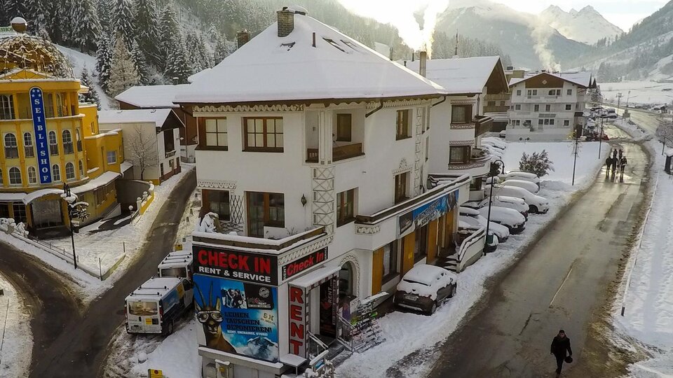 A charming building in the snow with a "CHECK IN" sign. In the background, there are snow-covered mountains and a quiet road.