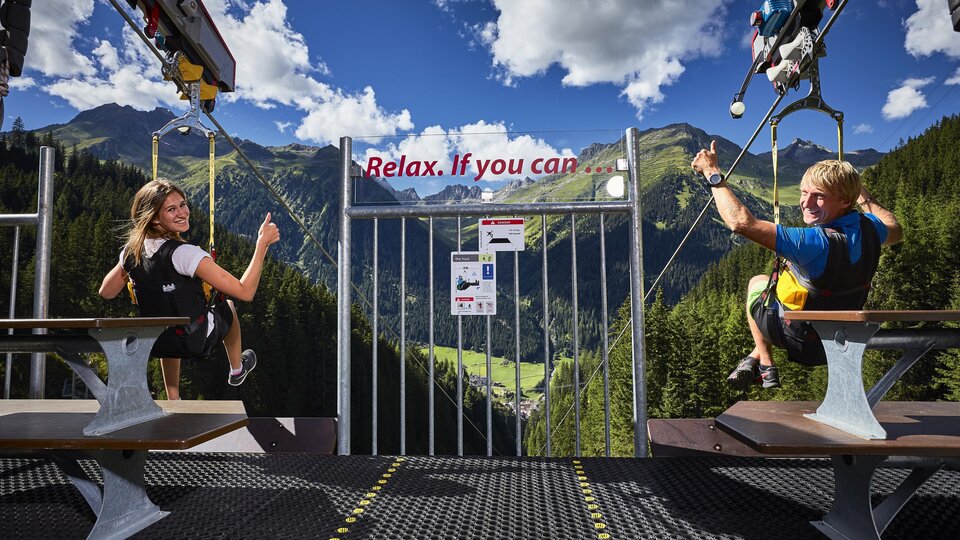 Zwei Personen sitzen auf einer Plattform mit Blick auf eine malerische Berglandschaft. Ein Schild fordert auf, sich zu entspannen.