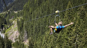 Eine Person schwebt an einer Seilrutsche über einen Wald aus hohen Tannen. Die Landschaft ist grün und bergig, ideal für Abenteuer in der Natur.