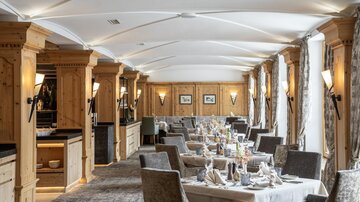 An elegant restaurant with wooden elements and cozy seating. The tables are festively set and large windows provide plenty of light.