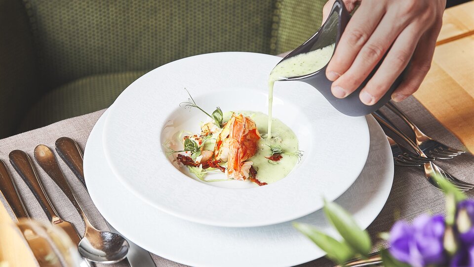 An elegant dish is served on a white plate. A hand pours a green sauce over the food, which is garnished with herbs.