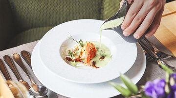 An elegant dish is served on a white plate. A hand pours a green sauce over the food, which is garnished with herbs.