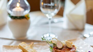 Ein elegantes Gericht auf einem weißen Teller, serviert mit frischen Beilagen. Im Hintergrund steht ein Glas Wein und ein Kerzenlicht.