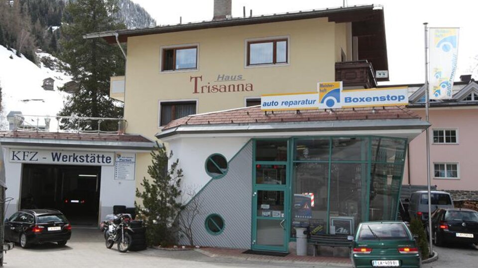 A cozy building with the sign "Haus Trisanna" and a workshop for car repairs. In the foreground, some cars and bicycles can be seen.