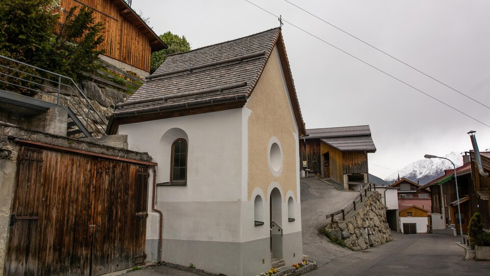 Eine kleine, weiße Kapelle steht an einer engen Straße, umgeben von traditionellen Holzhäusern. Der Himmel ist bewölkt und es scheint eine ruhige, ländliche Umgebung zu sein. | © Bernhard Gruber – Photography